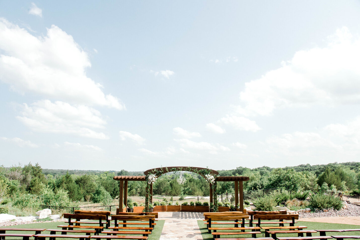 ECwedding158-copy-1450x968-1 - Stone Crest Venue Outdoor wedding ceremony setup with a wooden arch decorated with greenery and white flowers, facing rows of benches under a bright blue sky.